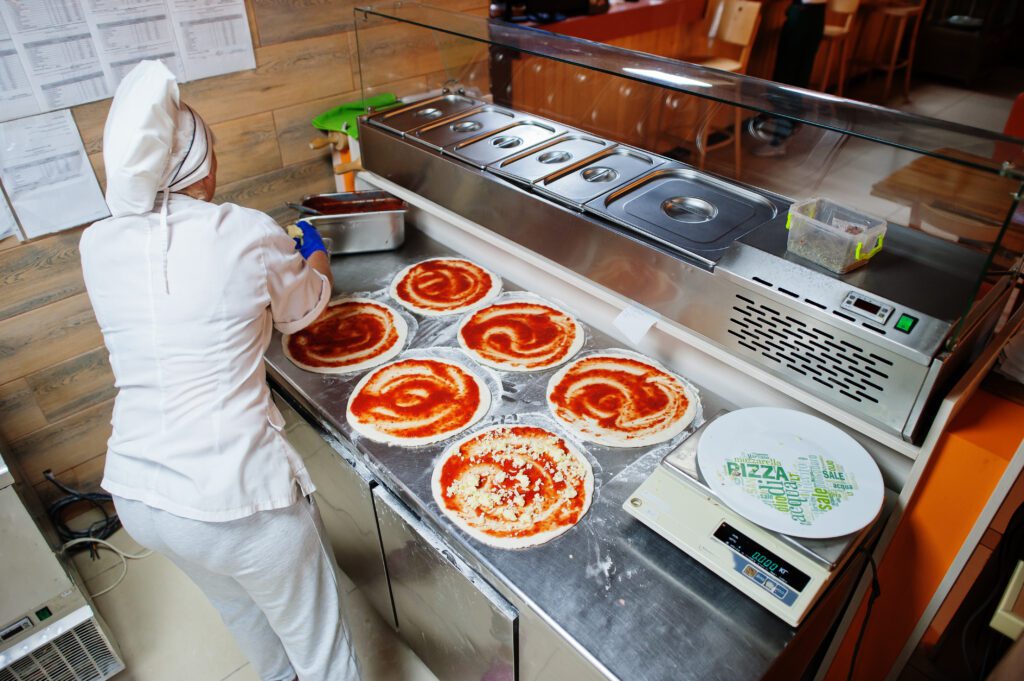 Professional pizza chef preparing multiple pizza bases with tomato sauce in a modern pizzeria kitchen, showcasing a stainless steel refrigerated prep table and ingredients setup for efficient pizza assembly.